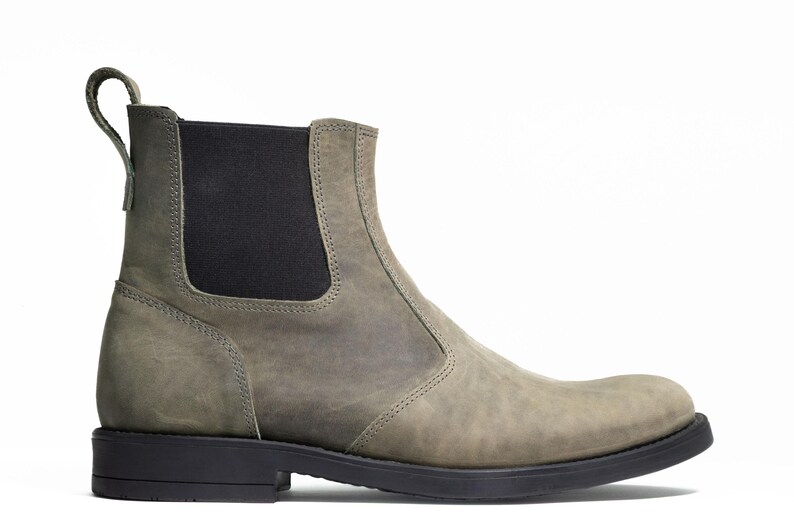Pode incluir: Vista lateral de uma bota Chelsea cinzenta com sola preta e painel lateral el&aacute;stico. A bota tem uma aba na parte de tr&aacute;s e &eacute; feita de um material texturizado semelhante ao couro. A bota tem aproximadamente 15 cm de altura.