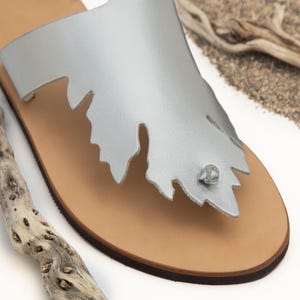 Puede incluir: Sandalias de cuero plateado con un dise&ntilde;o en forma de hoja y una suela de cuero marr&oacute;n.