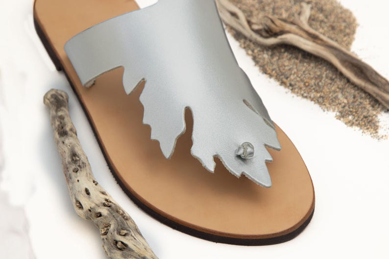 Puede incluir: Sandalias de cuero plateado con un dise&ntilde;o en forma de hoja y una suela de cuero marr&oacute;n.