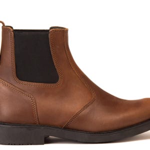 Pode incluir: Uma bota Chelsea de couro castanho com um painel lateral el&aacute;stico preto e uma presilha. A bota tem bico arredondado e sola preta. As costuras s&atilde;o vis&iacute;veis ao longo das costuras. A bota tem cerca de 15 cm de altura.