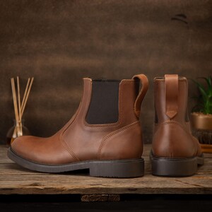 Pode incluir: Botas Chelsea de couro castanho com pain&eacute;is laterais el&aacute;sticos e puxadores. As botas t&ecirc;m sola castanha escura. S&atilde;o apresentadas numa superf&iacute;cie de madeira. Uma bota &eacute; vista de lado, a outra de tr&aacute;s.