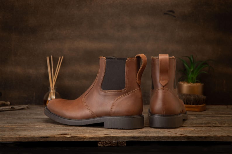 Pode incluir: Botas Chelsea de couro castanho com pain&eacute;is laterais el&aacute;sticos e puxadores. As botas t&ecirc;m sola castanha escura. S&atilde;o apresentadas numa superf&iacute;cie de madeira. Uma bota &eacute; vista de lado, a outra de tr&aacute;s.
