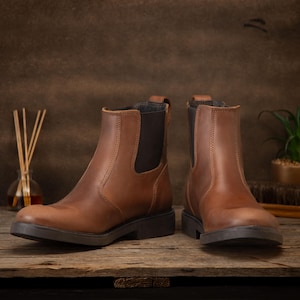 Pode incluir: Um par de botas Chelsea de couro castanho com pain&eacute;is laterais el&aacute;sticos e solas pretas. As botas s&atilde;o exibidas numa superf&iacute;cie de madeira, mostrando o seu design cl&aacute;ssico. O cal&ccedil;ado &eacute; adequado para v&aacute;rias ocasi&otilde;es. Altura: cerca de 20 cm.