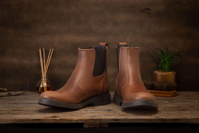 Pode incluir: Um par de botas Chelsea de couro castanho com pain&eacute;is laterais el&aacute;sticos e solas pretas. As botas s&atilde;o exibidas numa superf&iacute;cie de madeira, mostrando o seu design cl&aacute;ssico. O cal&ccedil;ado &eacute; adequado para v&aacute;rias ocasi&otilde;es. Altura: cerca de 20 cm.