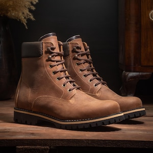 Puede incluir: Un par de botas de trabajo de cuero marrón con cordones marrones oscuros y suelas negras. Las botas tienen un aspecto robusto y desgastado y están sobre una superficie de madera. Cuello acolchado negro.