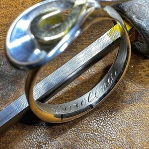 May include: A silver ring with an abstract design, featuring a curved, open shape. The ring is engraved with the name "Carolina" on the band. The ring rests on a brown leather surface, next to a metal tool and a wooden handle.