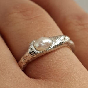 May include: A silver ring with a textured band and a small, oval pearl. The ring has a rustic, organic design, with a slightly irregular shape. The pearl is centered on the top of the ring, adding a touch of elegance.