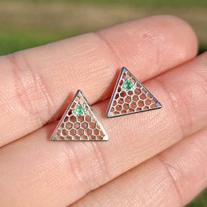 Può includere: Un paio di orecchini triangolari color argento. Ogni orecchino presenta un motivo a nido d'ape e una piccola gemma verde nella parte superiore. Gli orecchini sono esposti su una mano contro uno sfondo sfocato all'aperto.