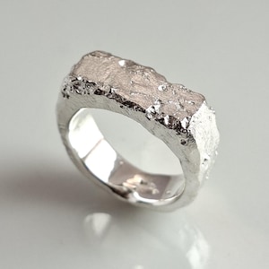 May include: A silver ring with a textured, irregular surface. The ring has a band and a raised, rectangular centerpiece with a rough, rocky appearance. The ring is set against a white background.