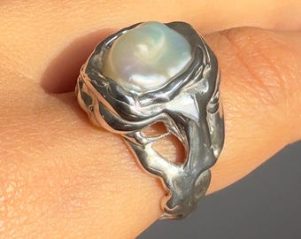 May include: A silver ring with an abstract, flowing design. It features a large, iridescent pearl. The metal has a textured, almost molten look, creating a unique piece of jewellery. The ring is a statement piece.