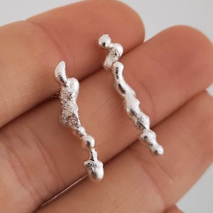 Puede incluir: Un par de pendientes de plata con un diseño único, orgánico y texturizado. Cada pendiente presenta una forma curva con múltiples pequeñas cuentas redondas.