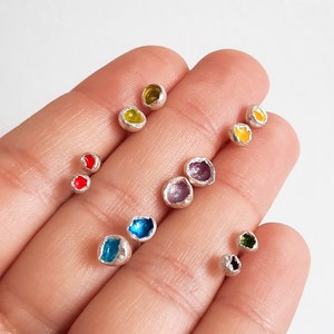 May include: A collection of small, silver stud earrings with colourful glass insets. The earrings are arranged in a hand, with the colours ranging from red, yellow, green, blue, and purple.