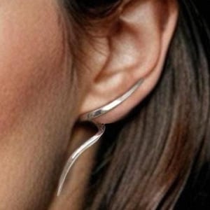 Könnte beinhalten: Ein silberner Ohrklemmen-Ohrring mit geschwungenem, abstraktem Design. Der Ohrring verläuft um das Ohr herum und erzeugt einen modernen, minimalistischen Look. Der Schmuck besteht aus glänzendem Metall.