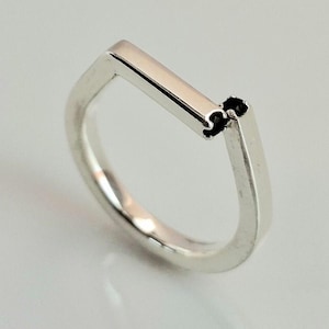 Op de afbeelding: Een minimalistische zilveren ring met een rechthoekig staafontwerp. De ring heeft een strakke, moderne uitstraling, met een gepolijste afwerking. De band heeft een eenvoudige, afgeronde vorm en de staaf staat loodrecht op de band.