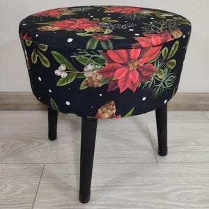 May include: A round upholstered stool with a black background and a floral pattern featuring red poinsettias, pinecones, and white berries. The stool has four black legs.