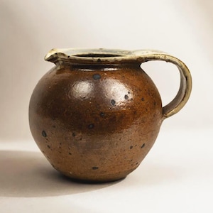 以下が含まれることがあります： 光沢のある茶色の釉薬に暗い斑点が全体に施された丸い陶器のピッチャー。この容器は幅広で膨らんだ本体、右側の湾曲したハンドル、上部の縁にある小さな注ぎ口が特徴で、縁は明るいクリーム色で仕上げられています。