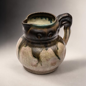 Può includere: Una piccola brocca in ceramica con corpo arrotondato e manico curvo. L'esterno presenta uno smalto maculato con toni marrone scuro e nero che colano su una base beige chiaro e rosa pallido. Il bordo interno mostra sfumature color ottanio.