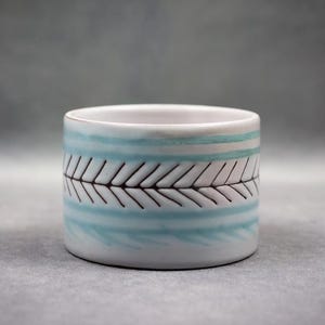 Peut inclure: Une petite tasse cylindrique en céramique avec un intérieur et un extérieur blancs. L'extérieur présente des bandes horizontales bleu clair et une bande centrale avec un motif noir en forme de feuille. La tasse est adaptée pour contenir de petits objets.