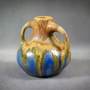Puede incluir: Un jarrón de cerámica con dos asas, con un cuerpo redondeado y un cuello estrecho. El jarrón está adornado con un esmalte en tonos azules, amarillos y marrones, creando una apariencia texturizada y artística. Las asas se curvan con gracia.