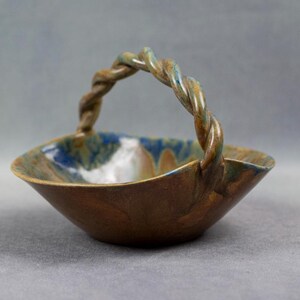 Puede incluir: Un pequeño cuenco de cerámica con un asa retorcida. El cuenco está esmaltado en tonos marrones, azules y verdes.