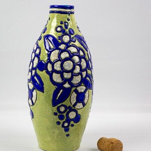 May include: A green ceramic bottle with blue and white floral designs. The bottle has a narrow neck and a cork stopper. The floral design includes large flowers and smaller circular accents.