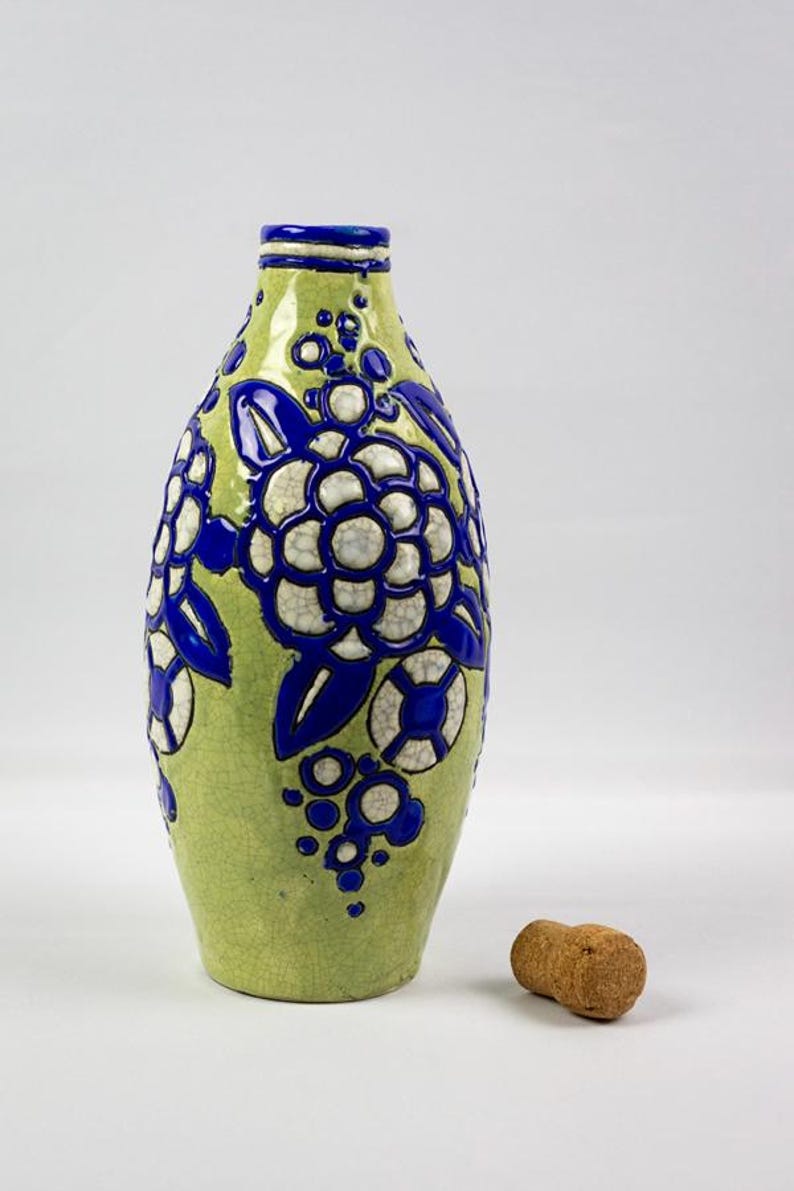 May include: A green ceramic bottle with blue and white floral designs. The bottle has a narrow neck and a cork stopper. The floral design includes large flowers and smaller circular accents.