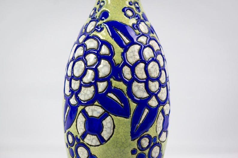 May include: A decorative ceramic vase with a bulbous shape. The vase features a light green background with a bold blue floral pattern. The flowers have a white, cracked texture, adding visual interest to the design.