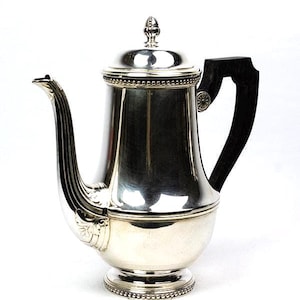 Puede incluir: Una cafetera plateada con un asa negra. La cafetera tiene un pico curvo y una tapa con un adorno decorativo. La base es redonda y se asienta sobre un pequeño pie decorativo. El asa es de madera oscura.