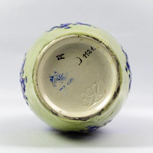 May include: A light green ceramic vase with blue floral accents. The bottom of the vase is visible, showing a cream-colored interior with stamped markings, including the number "892" and a blue stamp.