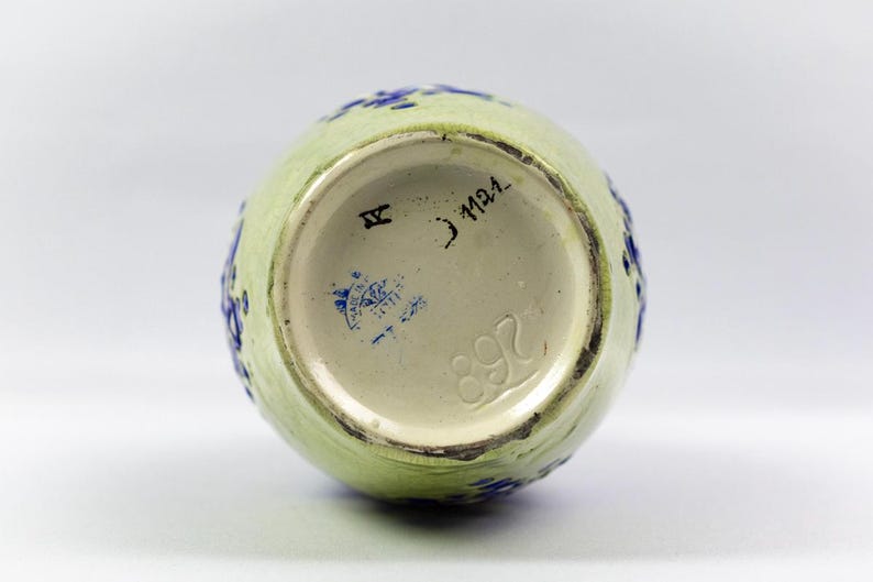 May include: A light green ceramic vase with blue floral accents. The bottom of the vase is visible, showing a cream-colored interior with stamped markings, including the number "892" and a blue stamp.