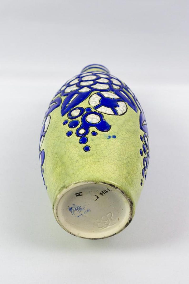 May include: A vintage ceramic vase with a light green background and blue floral designs. The vase has a textured surface and a round base. The bottom of the vase has markings including the number 892.