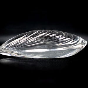 Puede incluir: Un objeto de vidrio transparente con forma de concha o almendra, que presenta una serie de crestas curvas grabadas en su superficie superior que captan la luz, creando una apariencia reflectante y suave sobre un fondo oscuro.