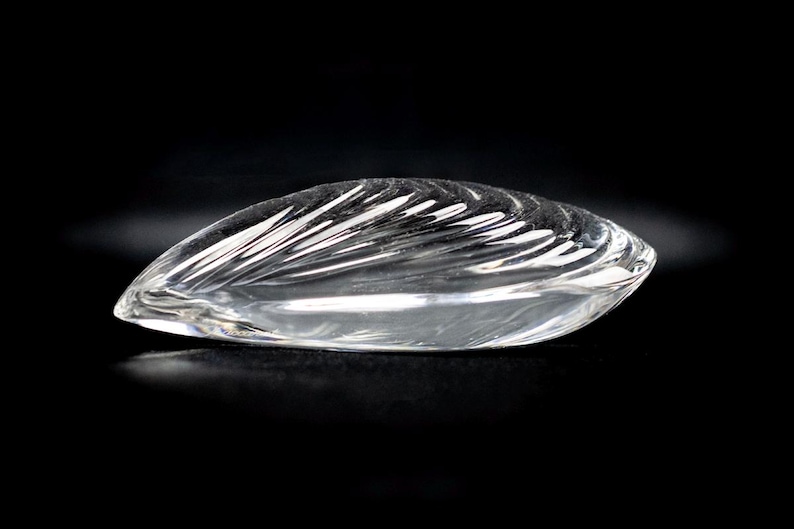 Puede incluir: Un objeto de vidrio transparente con forma de concha o almendra, que presenta una serie de crestas curvas grabadas en su superficie superior que captan la luz, creando una apariencia reflectante y suave sobre un fondo oscuro.