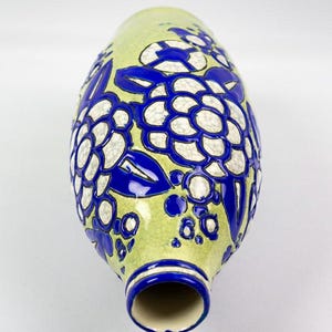 May include: A ceramic vase with a light green background and a floral design in white and blue. The vase has a narrow neck and a wide, rounded body. The floral pattern is repeated around the vase.