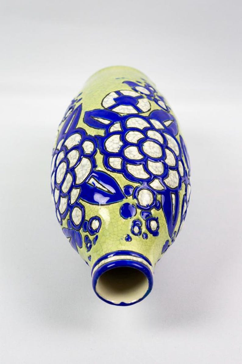 May include: A ceramic vase with a light green background and a floral design in white and blue. The vase has a narrow neck and a wide, rounded body. The floral pattern is repeated around the vase.