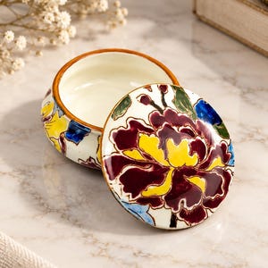 Pode incluir: Uma caixa de joias redonda em cerâmica com tampa removível, apresentando um design floral pintado à mão em bordô profundo, amarelo brilhante e azul sobre um fundo branco. O interior do recipiente é liso e creme com uma borda castanha.