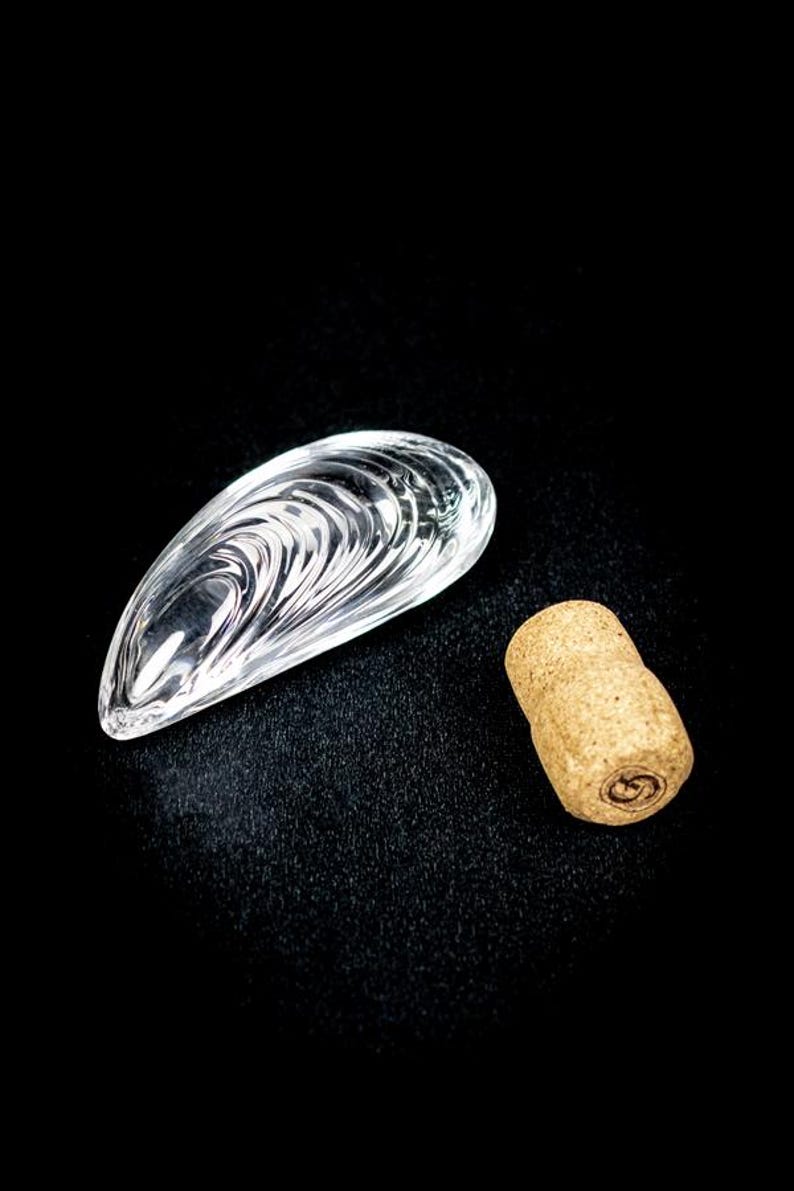 Puede incluir: Un objeto de vidrio transparente moldeado con forma de concha de mejill&oacute;n con crestas conc&eacute;ntricas visibles, colocado junto a un tap&oacute;n de corcho natural cil&iacute;ndrico de color marr&oacute;n claro con un peque&ntilde;o logotipo circular oscuro estampado.