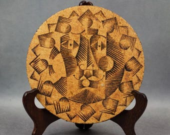 May include: A round ceramic plate with a brown and black geometric design. The design features a stylized face with a large nose and eyes.