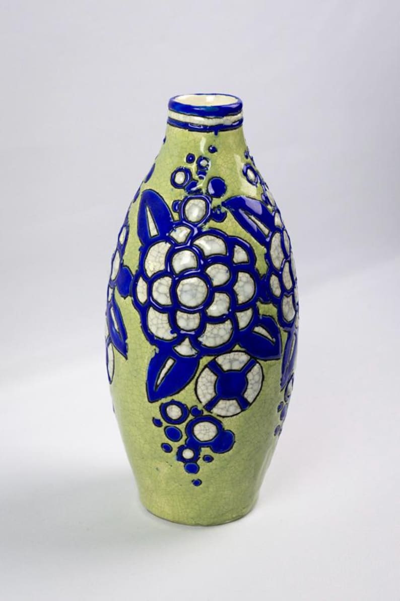 May include: A ceramic vase with a light green background and blue floral designs. The vase has a bulbous shape with a narrow neck and a blue rim. The floral pattern includes large flowers and smaller circular accents.