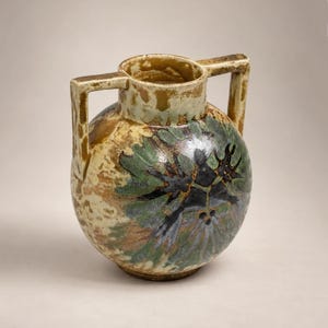 Peut inclure: Un vase en céramique rond doté de deux anses verticales sur les côtés. La surface présente un émail moucheté dans des tons crème, beige et marron, rehaussé par un motif abstrait floral ou en forme d'étoile vert foncé et noir sur le devant.