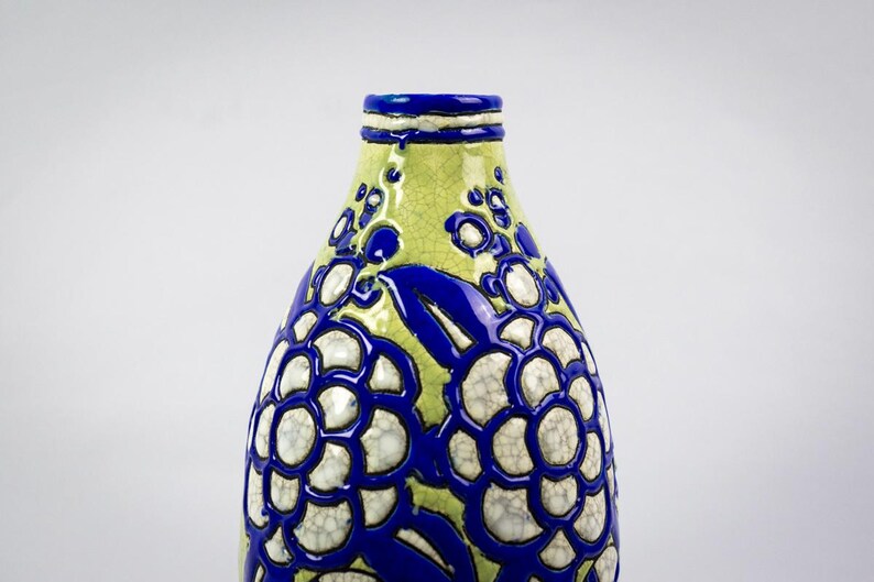 May include: A ceramic vase with a unique floral design. The vase features a light green background with a blue floral pattern. The flowers have a white center and a blue outline. The top and rim of the vase are blue.