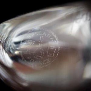 Puede incluir: Un objeto de vidrio transparente y curvo que presenta un sello circular grabado en su superficie. El sello contiene el texto "CRISTAL" en la parte superior, "D'ARQUES" en el centro y "FRANCE" en la parte inferior, en una fuente sans-serif.