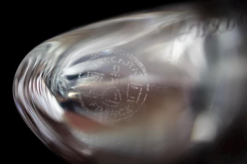 Puede incluir: Un objeto de vidrio transparente y curvo que presenta un sello circular grabado en su superficie. El sello contiene el texto "CRISTAL" en la parte superior, "D'ARQUES" en el centro y "FRANCE" en la parte inferior, en una fuente sans-serif.