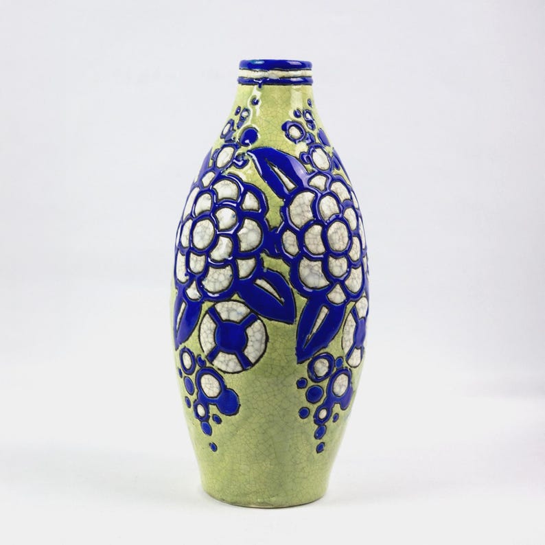 May include: A tall, decorative vase with a light green background and a blue and white floral design. The vase has a narrow neck and a rounded body. The floral pattern is repeated on both sides of the vase, creating a symmetrical design.
