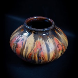 May include: A ceramic vase with a rounded body and a wide opening. The vase is decorated with a colourful glaze that features shades of brown, orange, and yellow.