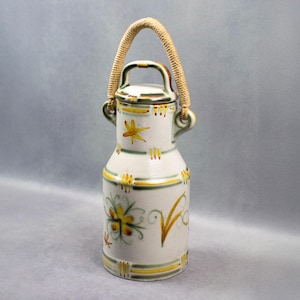 Puede incluir: Una jarra de leche de cerámica blanca con un diseño floral amarillo y verde y un asa de cuerda. La jarra tiene una tapa y está decorada con una estrella y rayas.