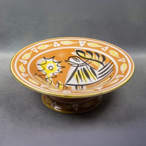 Peut inclure: Un bol en céramique avec un motif d'oiseau stylisé en noir et jaune sur fond orange. Le bol a un pied et un bord jaune.