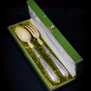 Peut inclure: Un ensemble de couverts anciens, comprenant une cuillère et une fourchette, présenté dans un coffret vert doublé de velours. La cuillère et la fourchette ont des têtes de couleur ivoire et des manches argentés ornés. La boîte est verte avec un fermoir doré.
