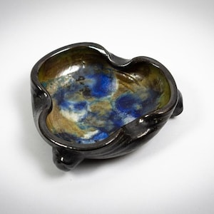 Peut inclure: Un petit plat en céramique noire avec trois pieds. L'intérieur du plat est émaillé dans des tons de bleu, vert et marron.