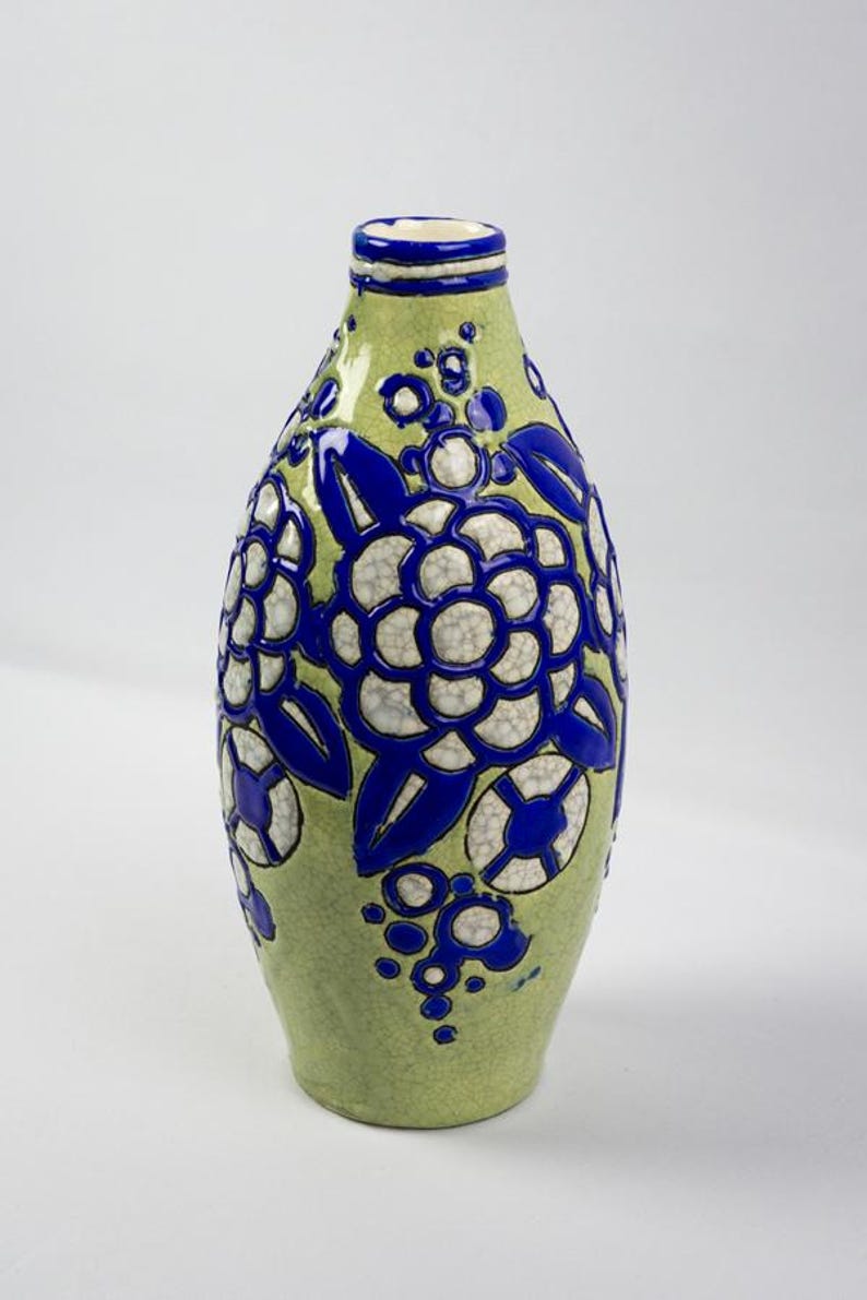 May include: A green ceramic vase with a floral design. The vase has a rounded body and a narrow neck. The floral pattern is white and blue, with the flowers and leaves outlined in blue. The vase is approximately 10 inches tall.